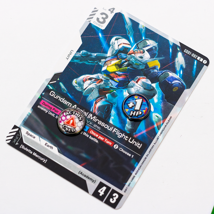 Gundam Photo 3 Trading card featuring 'Gundam Aerial (Mirasoul Flight Unit)' from the Gundam TCG. The card showcases a dynamic illustration of the mecha with glowing red accents. Two Battle Baubles tokens - '+1 HP' and 'ACTION USED' - are placed on the card to indicate active effects.